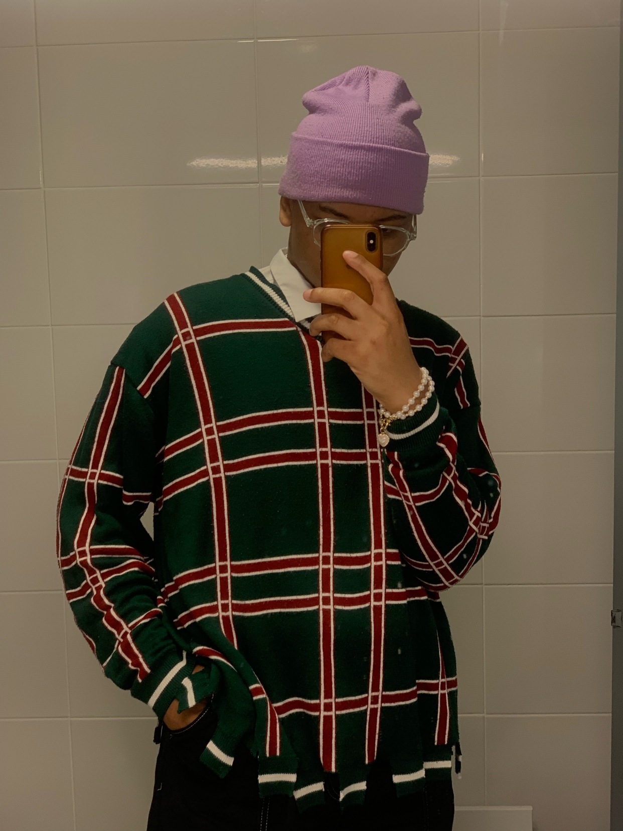 A person wearing a light purple beanie, clear glasses, and a green and red plaid sweater over a white collared shirt takes a mirror selfie. They are also wearing a pearl bracelet on their left wrist.