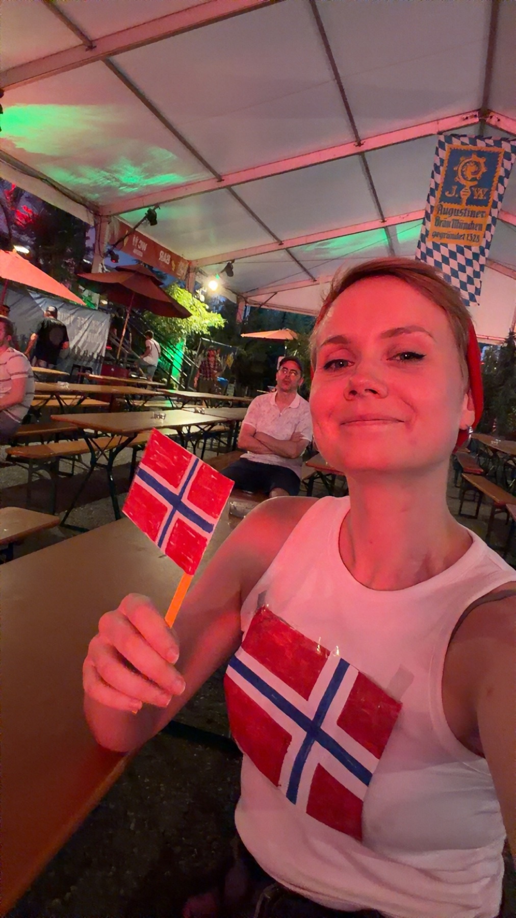 A woman smiles at the camera while holding two small Norwegian flags. She is wearing a white tank top and a red headband. The background appears to be an outdoor event under a tent with tables and other people.