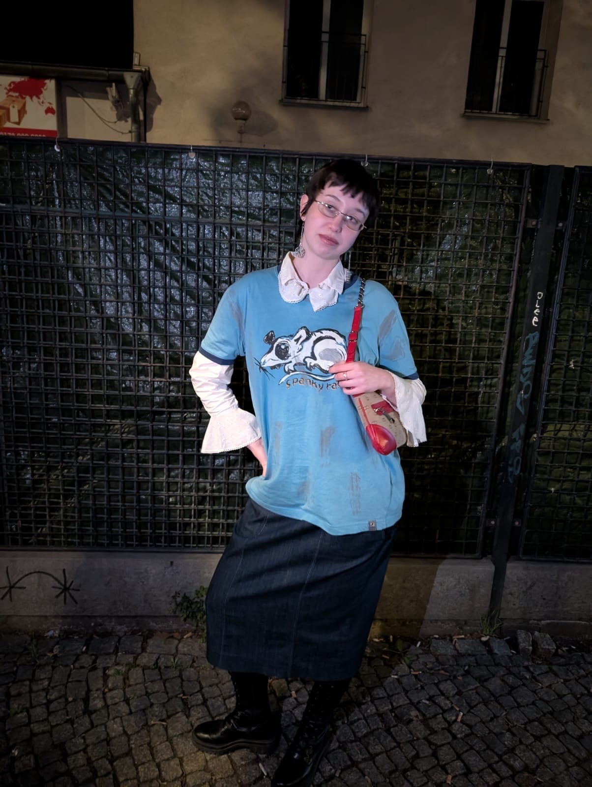 A woman with short hair and glasses poses in a blue t-shirt with a rat graphic over a white collared shirt, paired with a denim skirt and black boots. She carries a small bag over her shoulder.