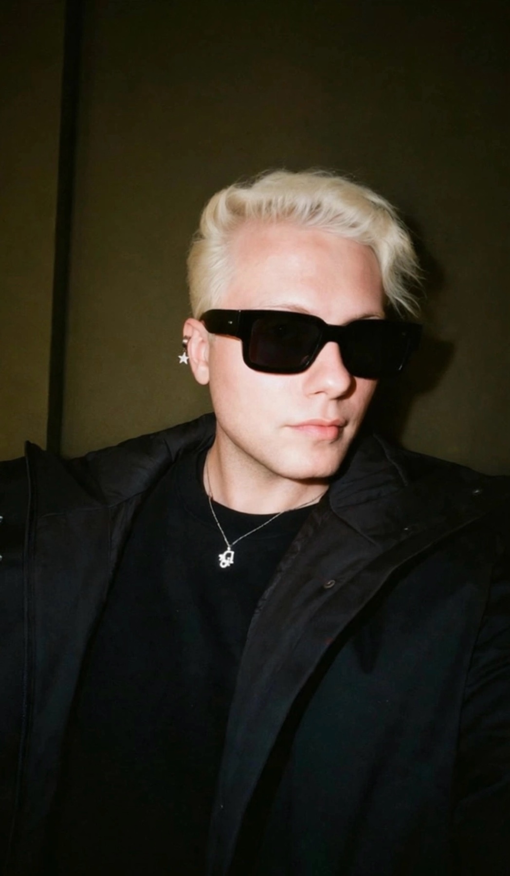 A man with blonde hair and sunglasses poses for a selfie. He is wearing a black t-shirt and a black jacket, with a necklace and earrings visible.
