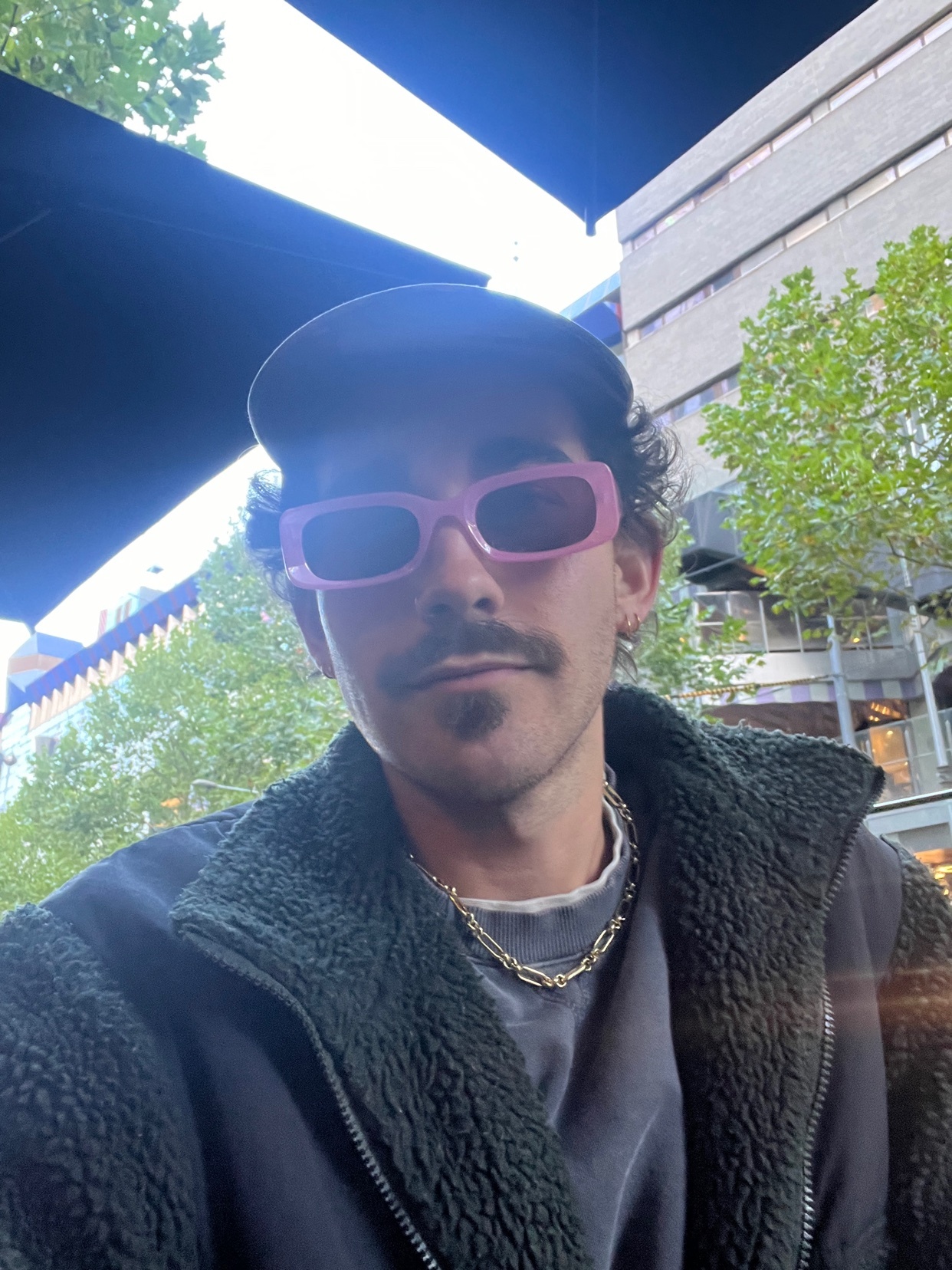A man wearing pink sunglasses, a baseball cap, and a chain necklace poses for a selfie outdoors.