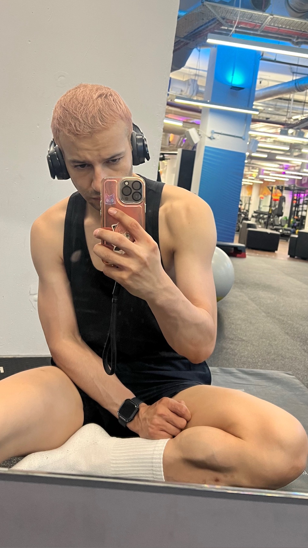 A man with pink hair takes a selfie in a gym while wearing headphones and a black tank top. He is sitting down and appears to be flexing his arm.