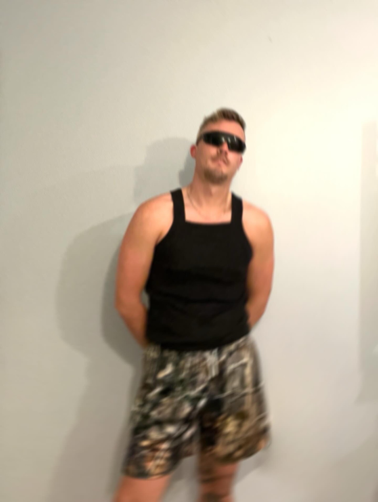 A man poses in a black tank top and camouflage shorts, wearing sunglasses. The photo has a slightly blurred effect.