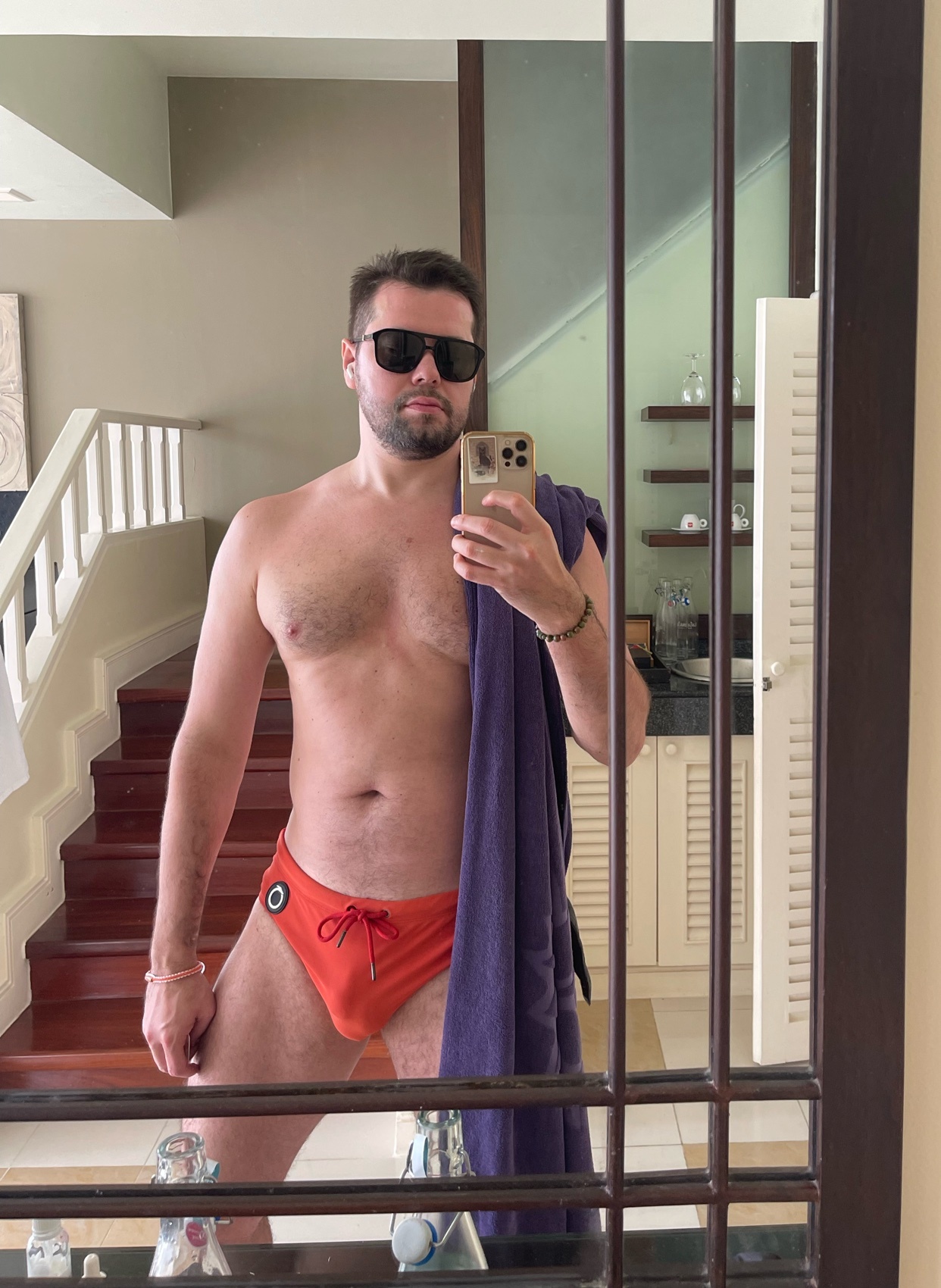 A man takes a mirror selfie, shirtless and wearing orange swimwear. He is wearing sunglasses and holding a towel.