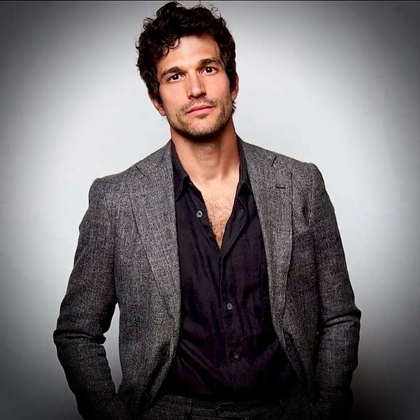 A man in a gray suit and black shirt poses for a photo. He has dark, curly hair and a confident expression.