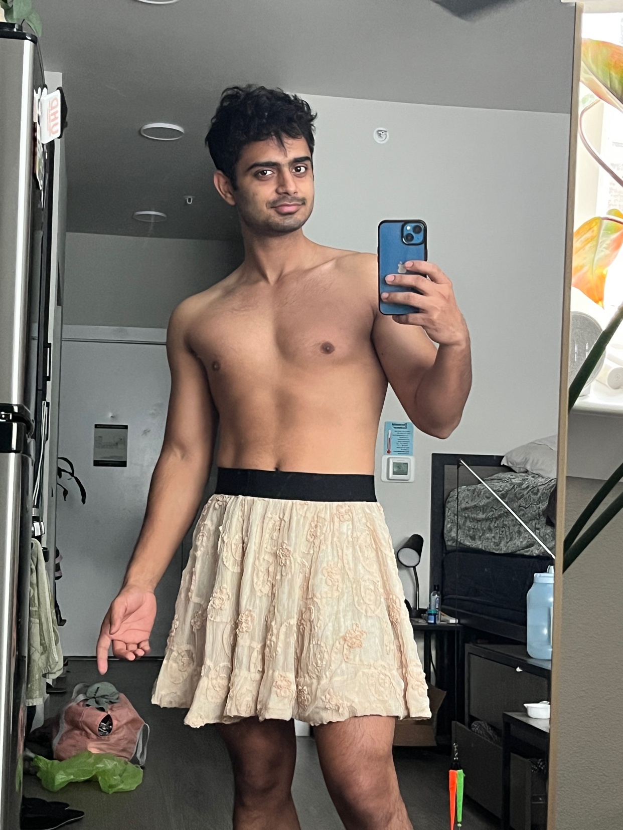 A man poses shirtless in a skirt, taking a mirror selfie. He is flexing his body.