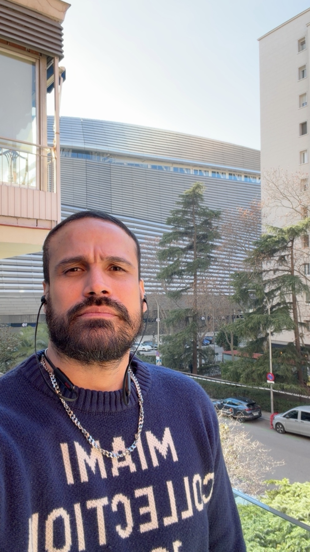 A man takes a selfie outdoors in Madrid. He is wearing a sweater and has a beard.