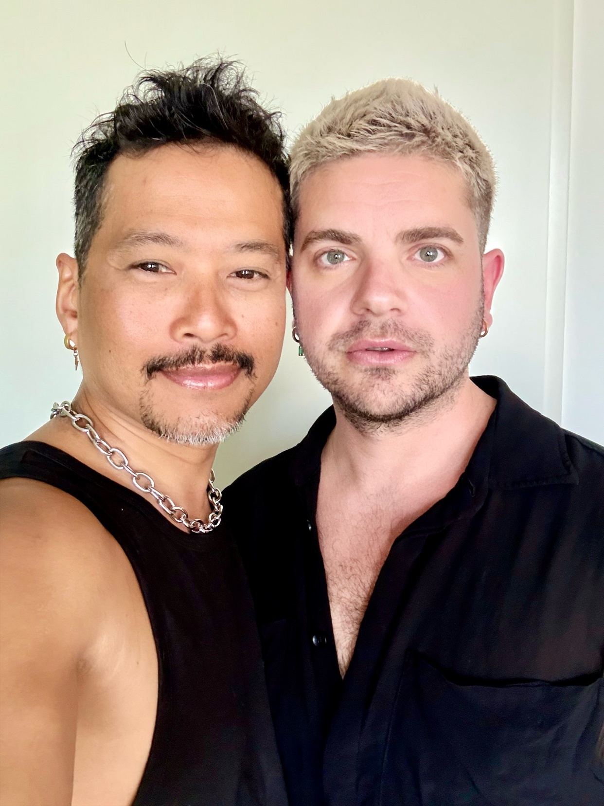 A close-up, waist-up photo shows two men posing together. The man on the left has tan skin, a mustache, and graying stubble, wearing a black tank top and a silver chain. The man on the right has fair skin, blond hair, and stubble, wearing a black but