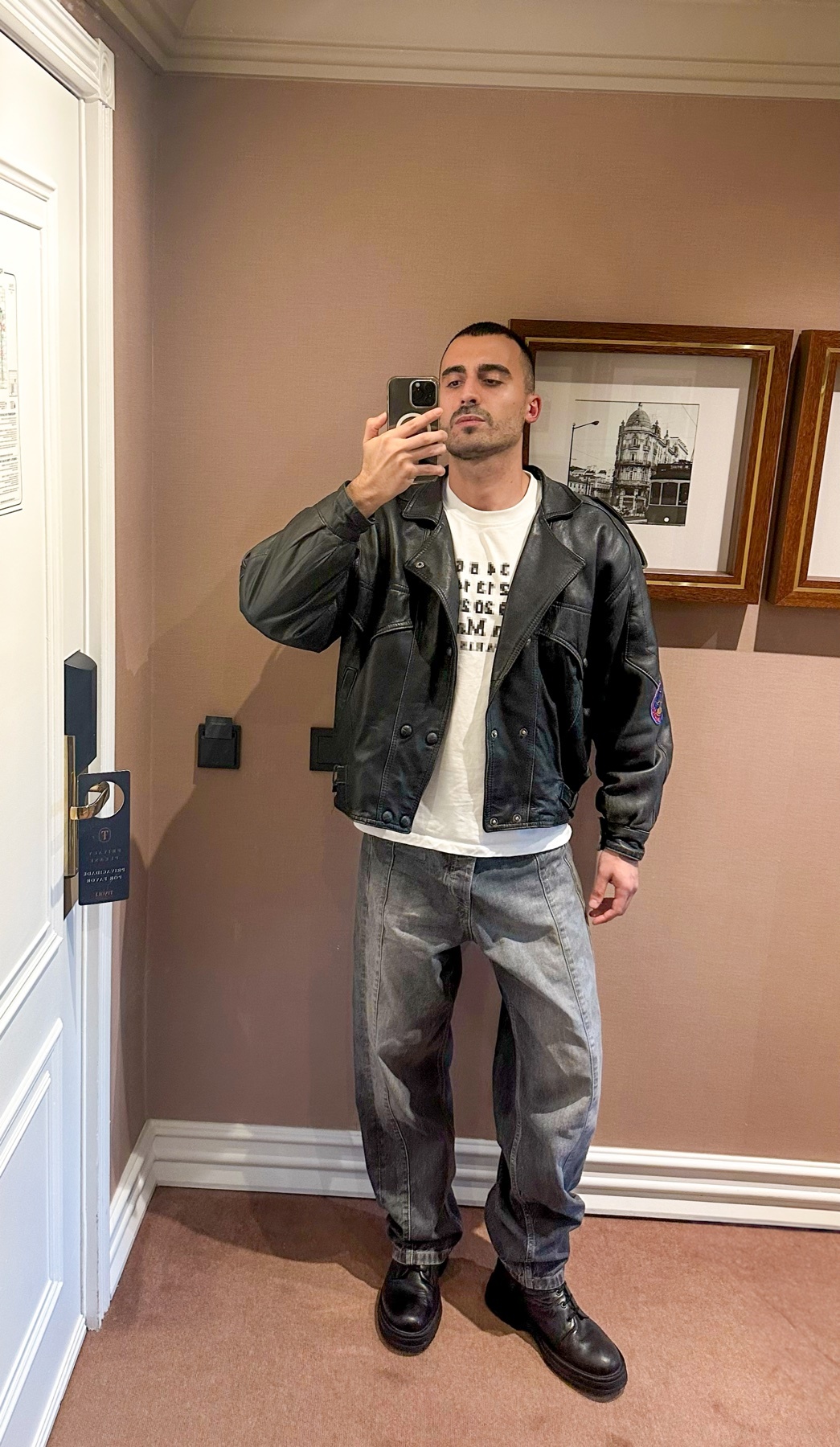 A man takes a selfie in a hotel room, wearing a leather jacket over a graphic t-shirt and baggy jeans.
