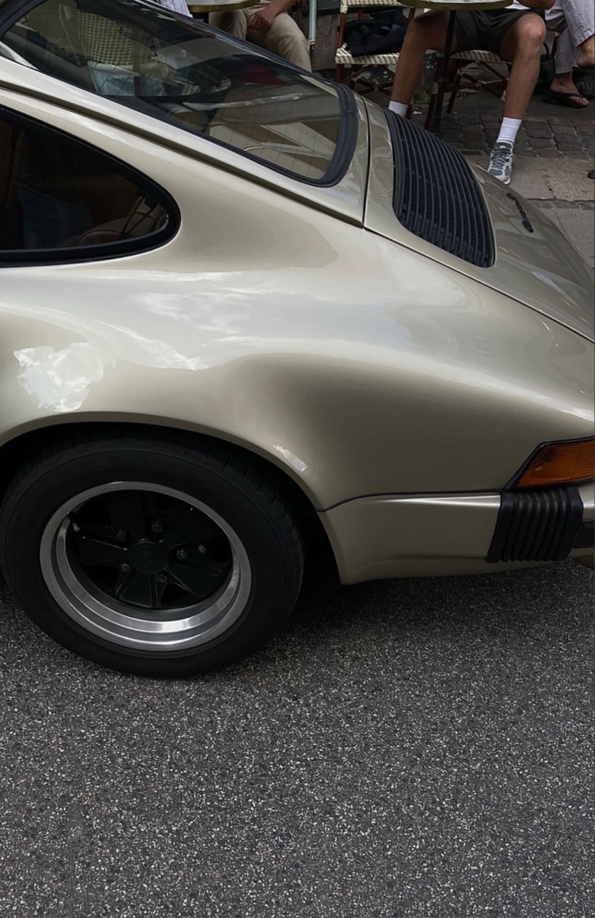 A close-up shot of a classic silver Porsche. The car is parked on a street, with a glimpse of people in the background.