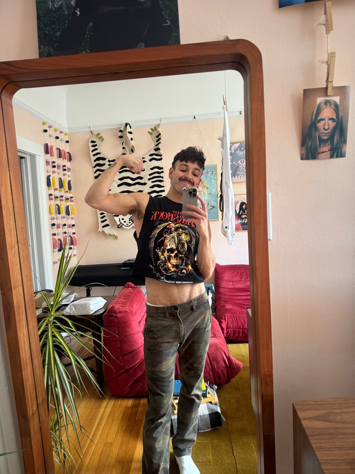 A man with a mustache and a piercing poses in front of a mirror, flexing his bicep. He is wearing a cropped graphic t-shirt and camouflage pants.