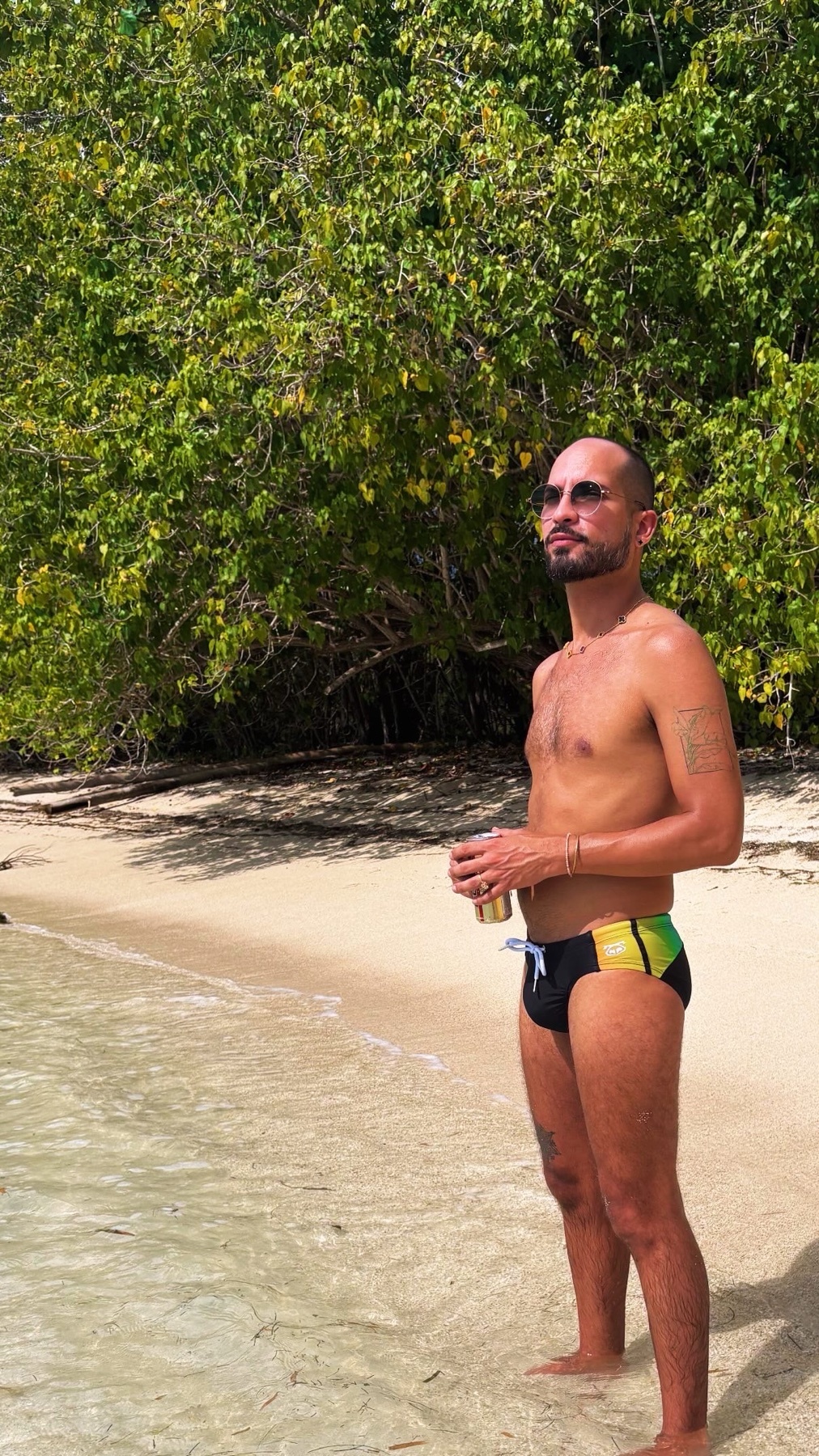 A man stands on a sandy beach wearing colorful swimwear and sunglasses. He holds a drink in his hand and has tattoos on his arms.