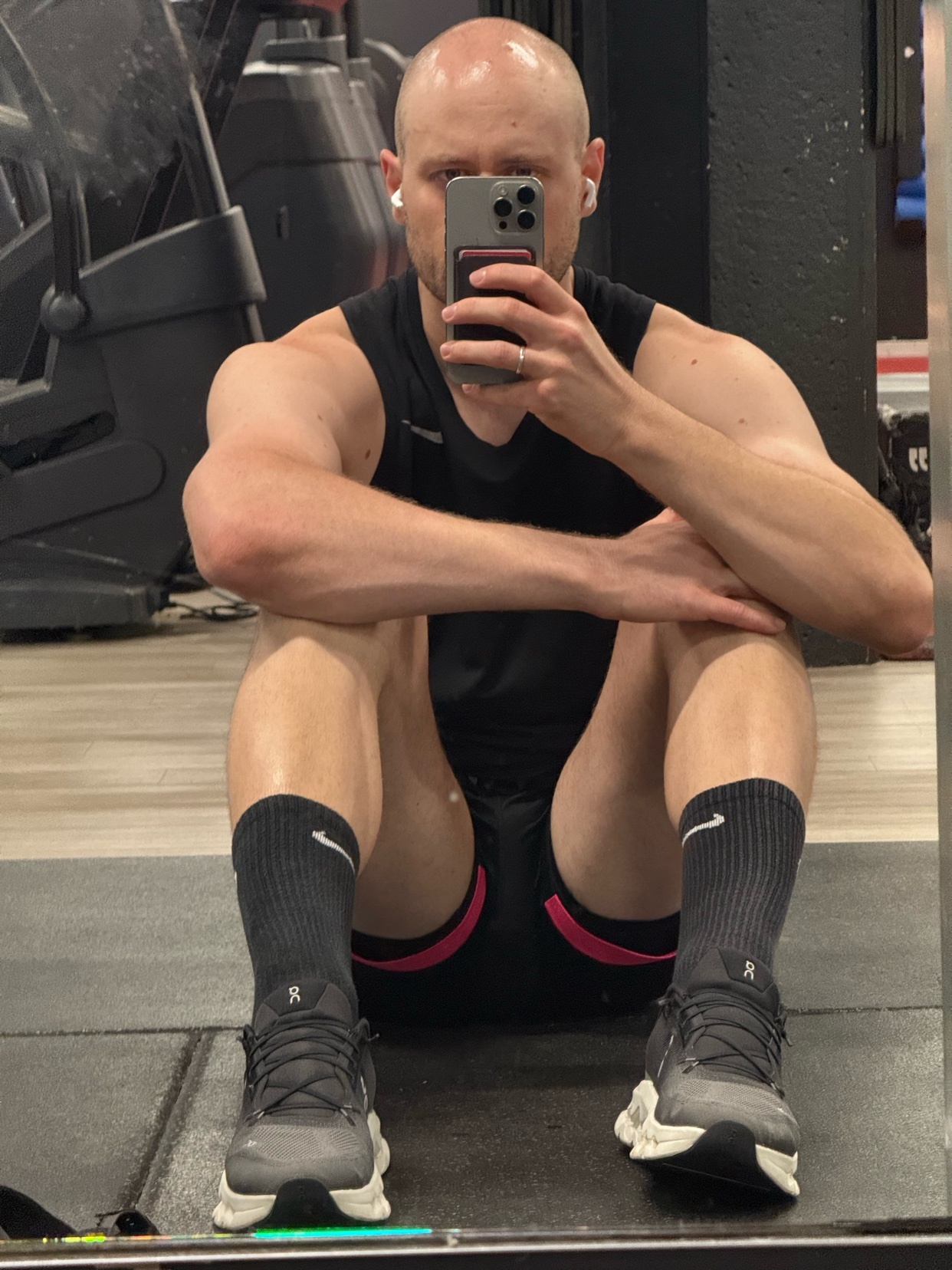 A bald man in athletic wear takes a mirror selfie while sitting on the floor of a gym. He is wearing a black tank top, black shorts with pink trim, and grey sneakers.