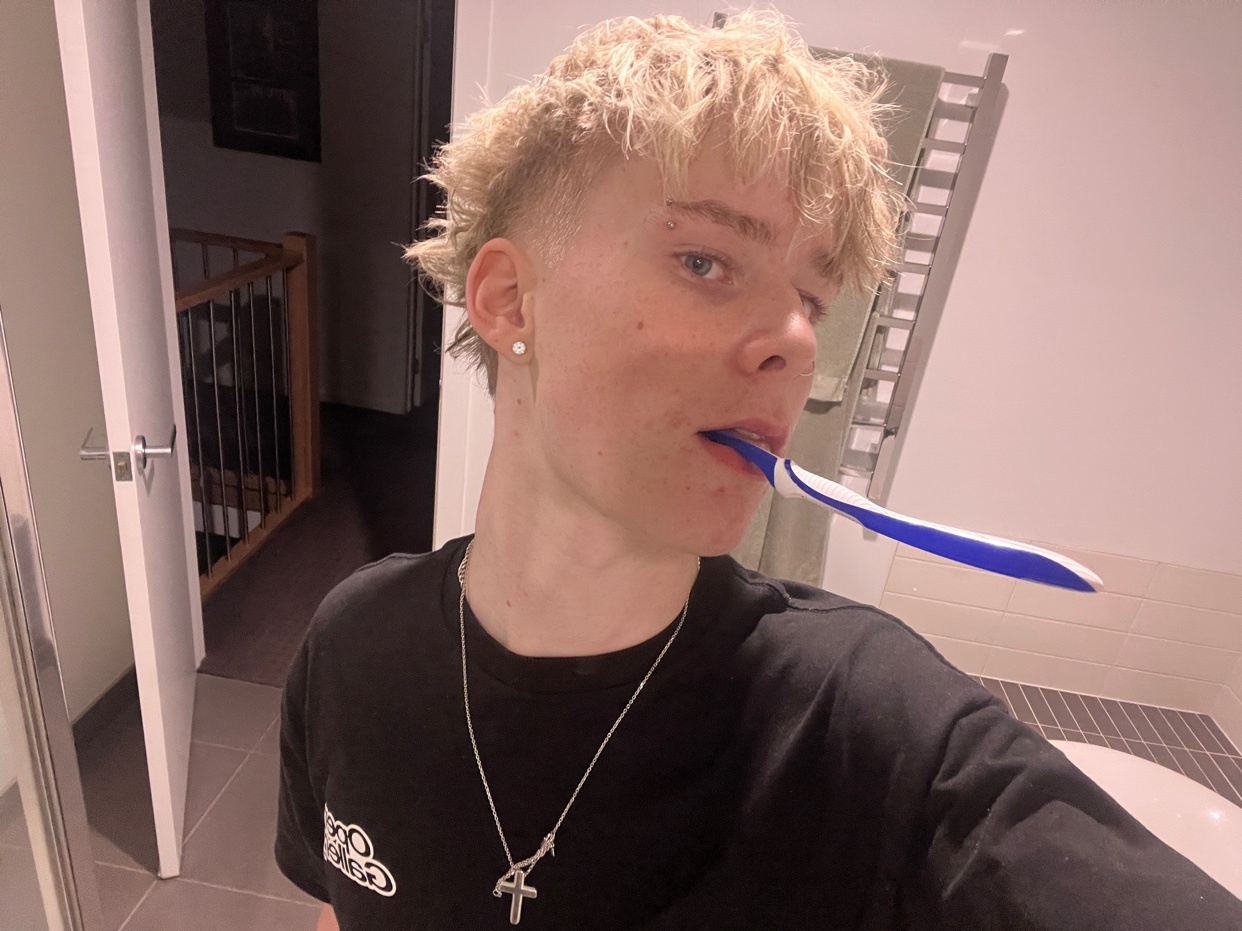 A teen boy with short, spiky blond hair and a mullet poses for a selfie in a bathroom while holding a toothbrush in his mouth.