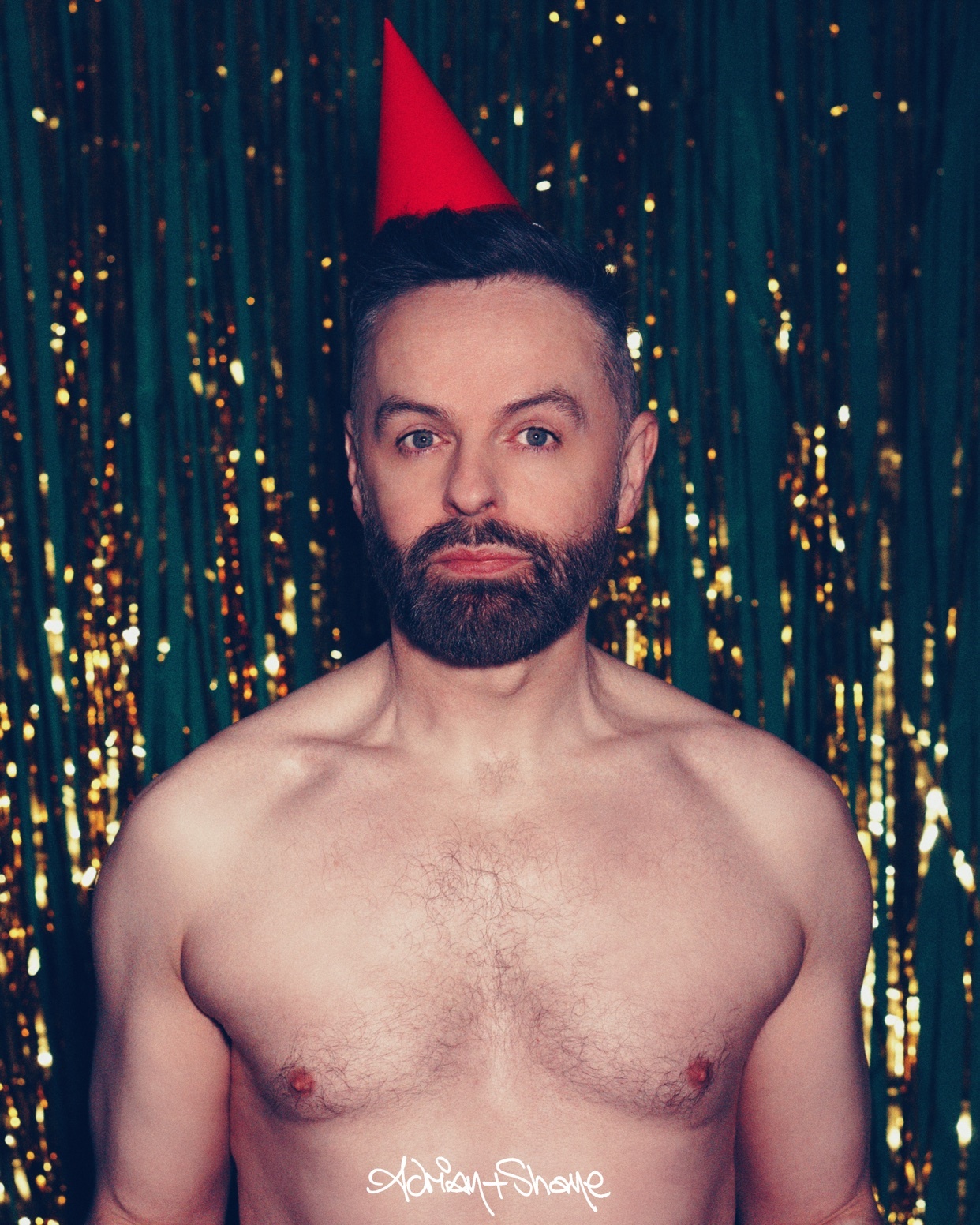 A man with a beard and a red party hat is shirtless against a backdrop of green and gold tinsel.