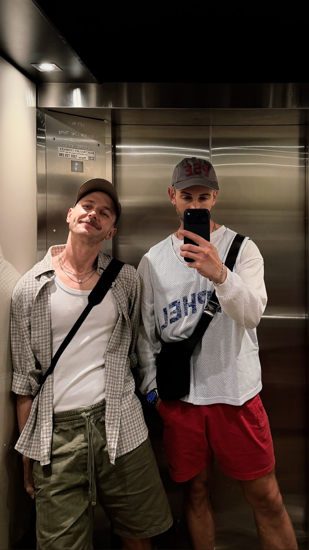 Two men pose for a selfie in an elevator, dressed in casual streetwear. The man on the left wears a plaid shirt over a tank top and shorts, while the man on the right wears a jersey over a long-sleeved shirt and red shorts.