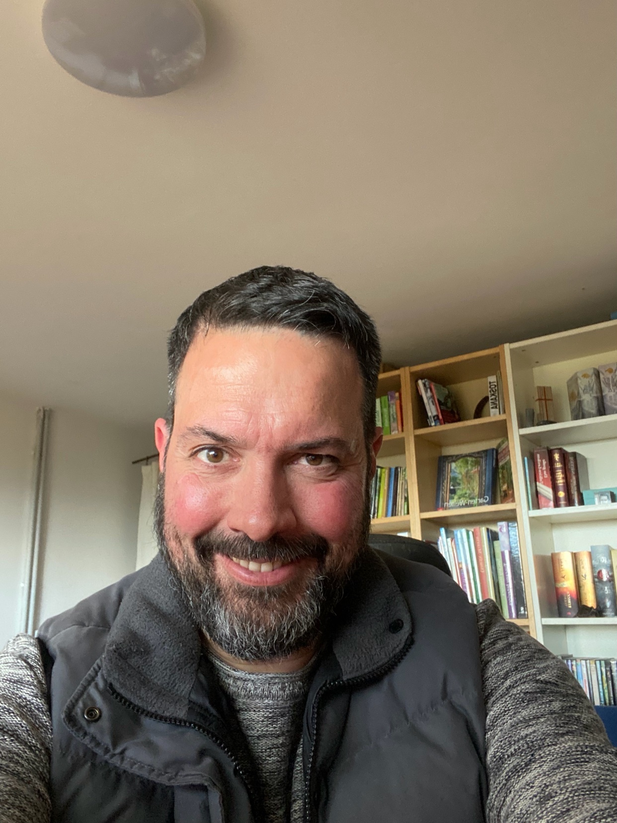 A man with a beard smiles at the camera, wearing a vest over a sweater. Behind him, a bookshelf is filled with books.