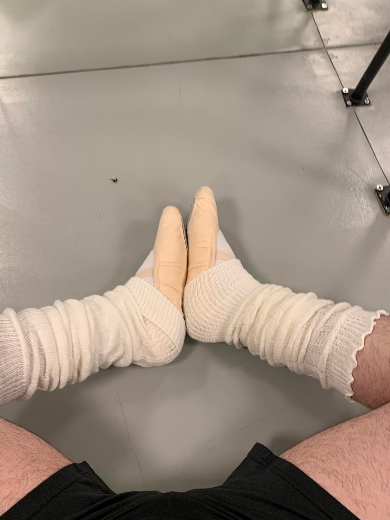 A close-up shot of a man's feet in ballet shoes and leg warmers. The image is taken from a low angle, emphasizing the feet and lower legs.