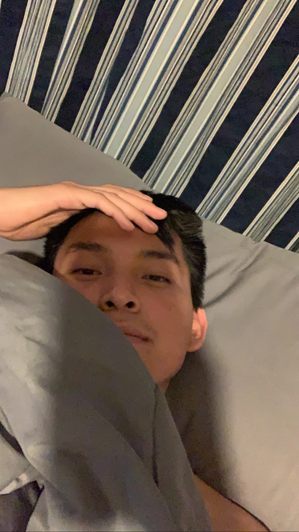 A man is lying in bed, looking at the camera. He is holding his hand on his head.