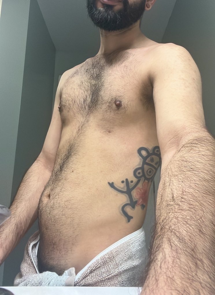 A man poses shirtless, revealing a tattoo on his side. The photo is a selfie taken in a bathroom.