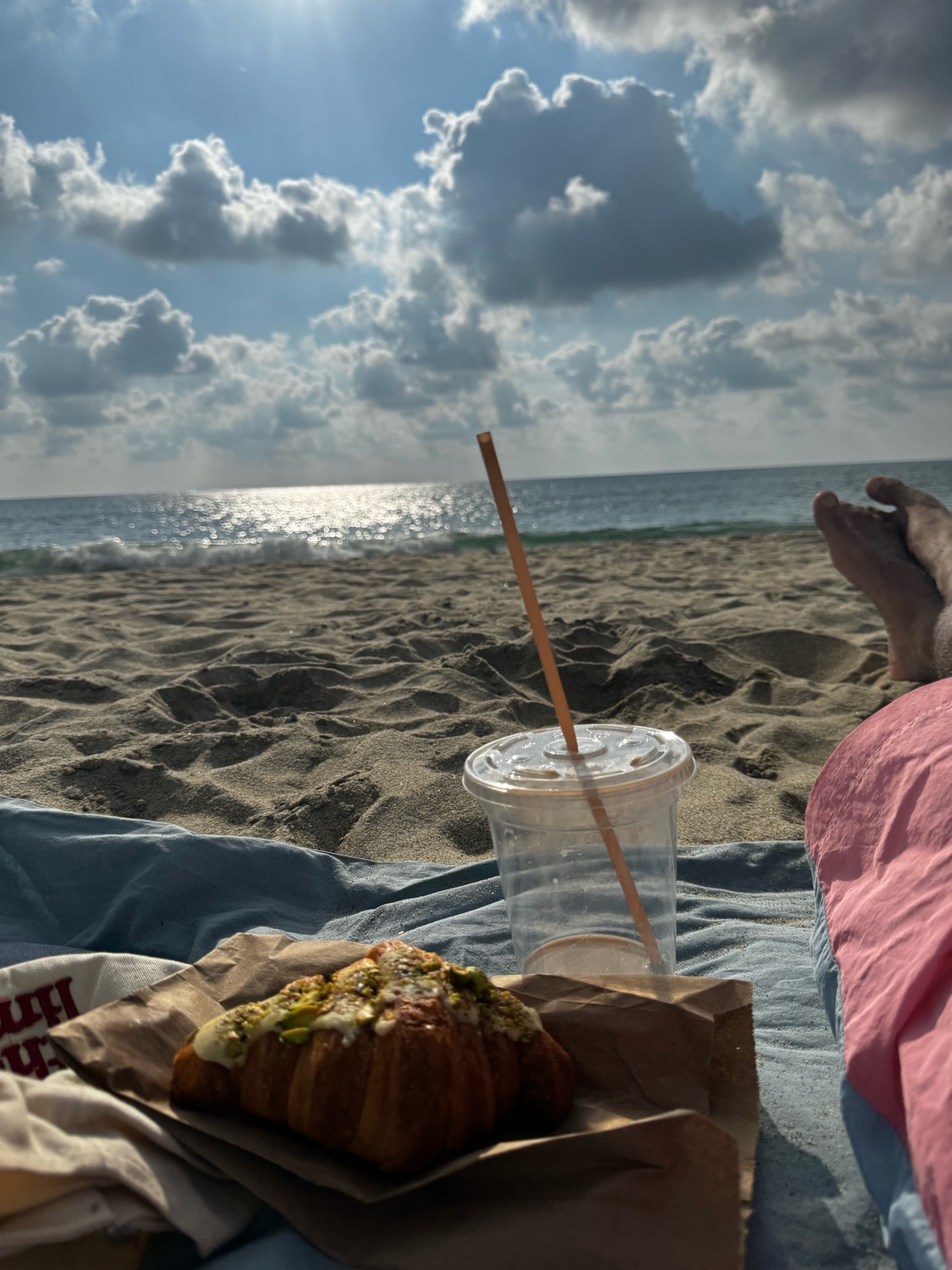 A close-up shot of a croissant and a drink on a beach blanket, with the ocean and sky in the background. The sun is shining brightly, casting a warm glow on the scene.