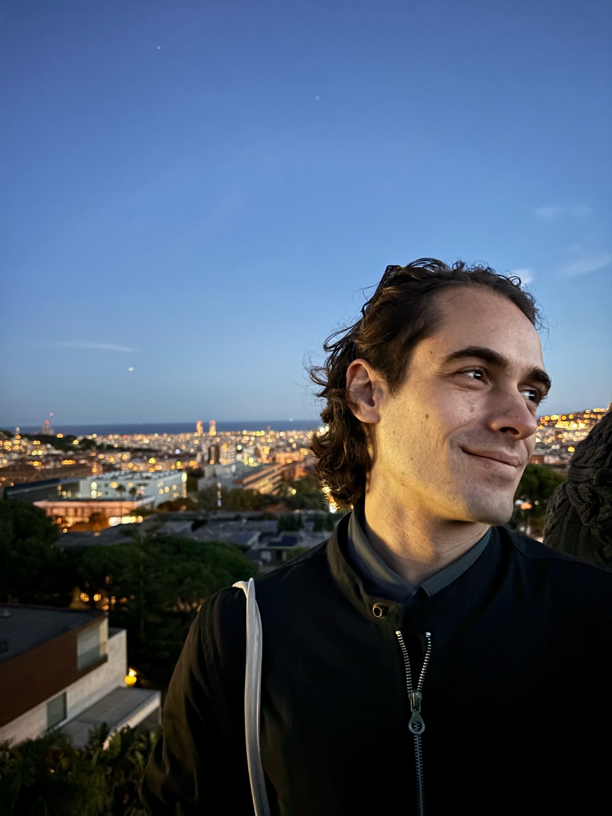 A man poses with a cityscape in the background. The photo is taken at night, with the city lights illuminating the scene.