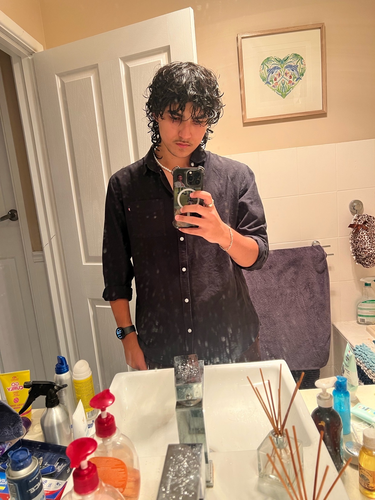 A man takes a mirror selfie in a bathroom. He is wearing a black shirt and has curly dark hair.