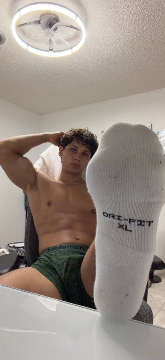 A shirtless man with curly dark hair poses with his foot forward, wearing green underwear and white socks. He is seated in a chair with his hand behind his head, showcasing his muscular physique.
