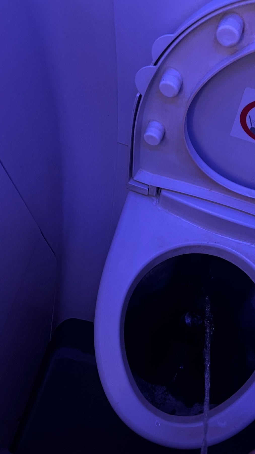 A close-up shot of a toilet with the lid up, illuminated by blue light. Water is running into the bowl.