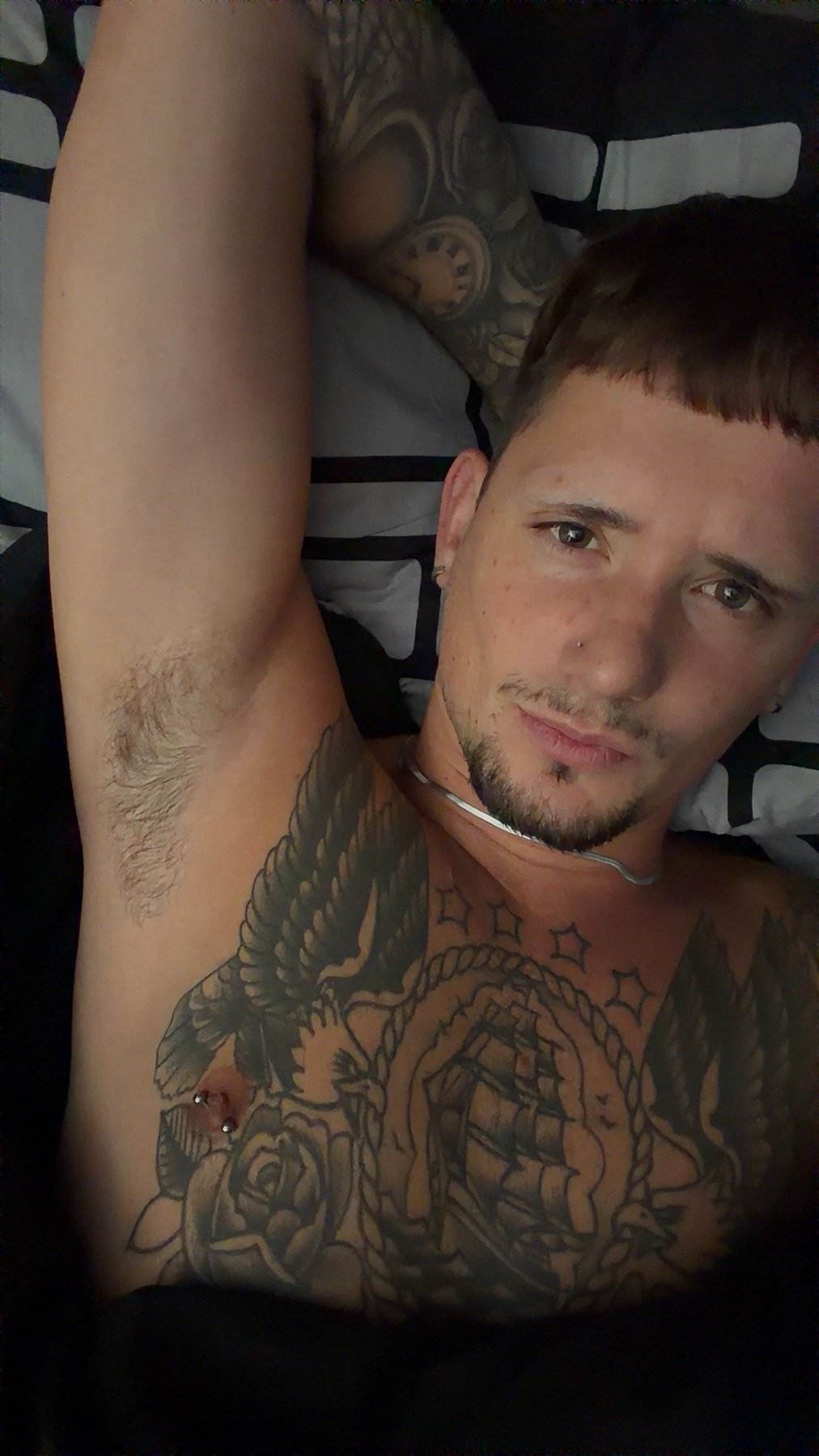 A man with extensive tattoos on his chest and arm poses for a close-up selfie. He has short, dark hair and stubble on his face.