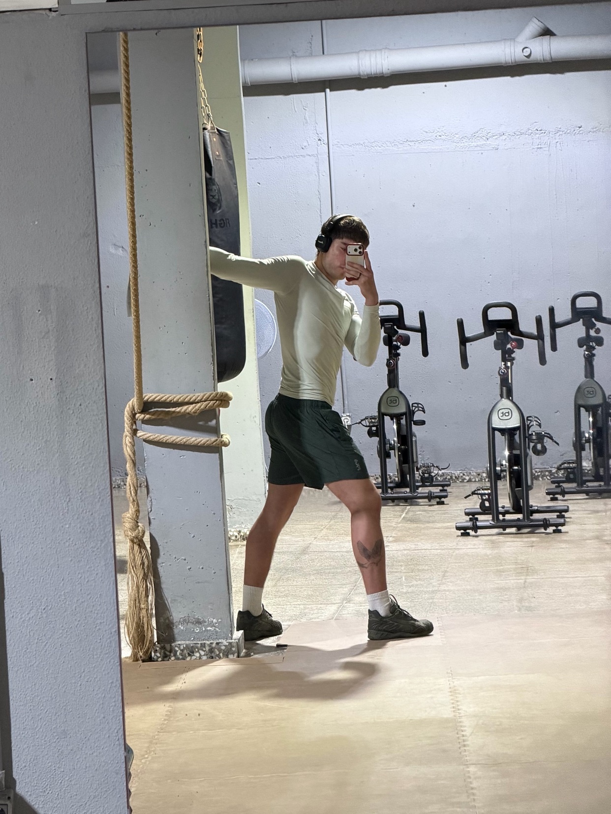A man takes a mirror selfie in a gym, wearing headphones and athletic wear. He is standing next to a punching bag with exercise bikes in the background.