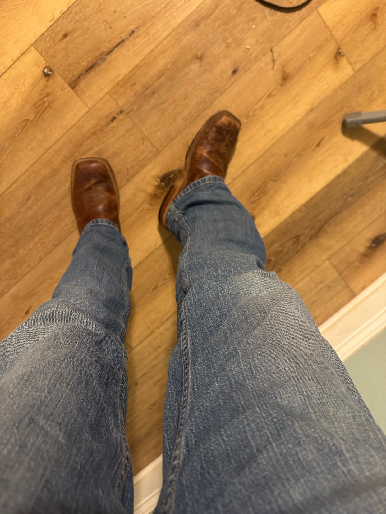 A person wearing blue jeans and brown cowboy boots stands on a wooden floor. The boots are the focal point of the image.