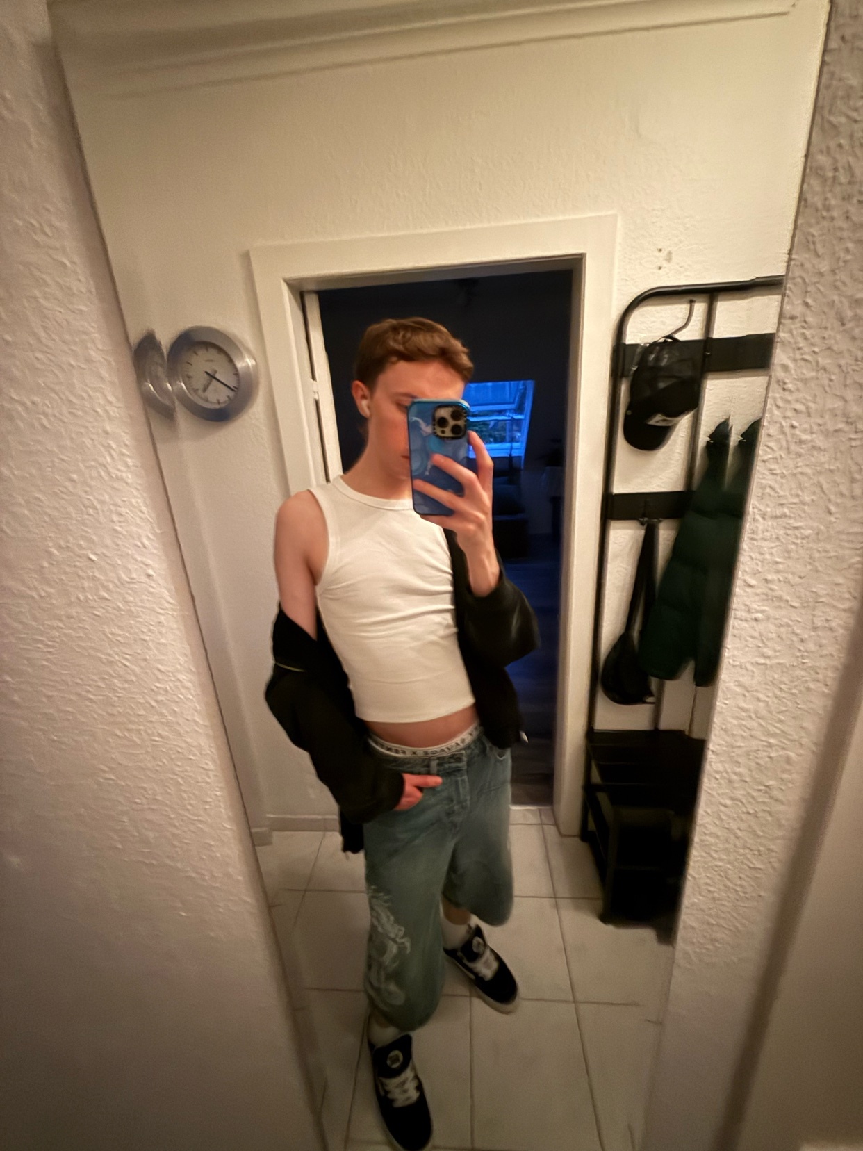 A man takes a mirror selfie, showcasing his outfit. He is shirtless, wearing jeans and sneakers.