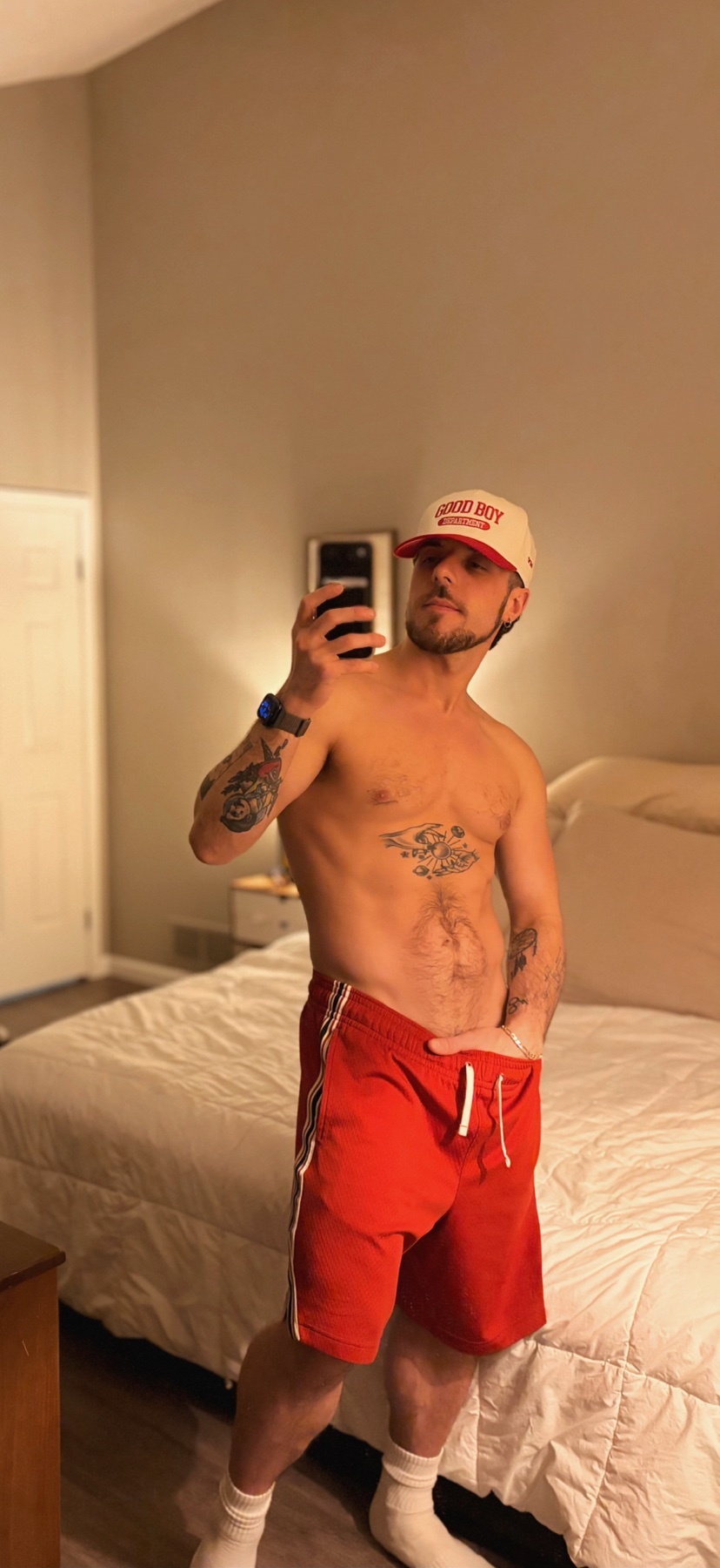 A man takes a mirror selfie, shirtless and wearing red shorts and a cap. He is in a bedroom, showing off his physique.