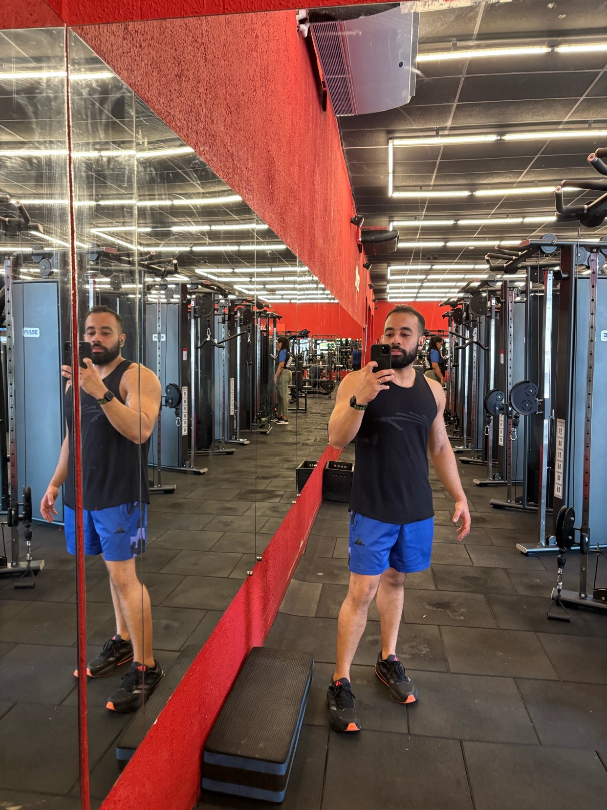 A man takes a mirror selfie in a gym, wearing a black tank top and blue shorts. The reflection shows gym equipment in the background.