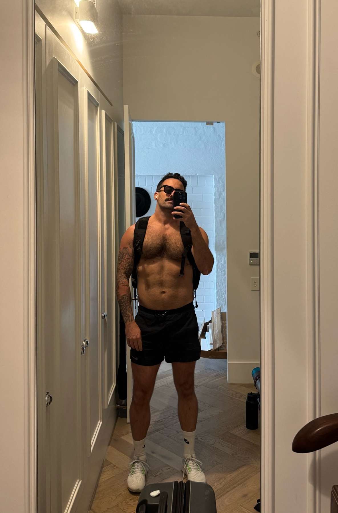 A man poses shirtless in front of a mirror, showcasing his physique. He wears shorts, sunglasses, and a backpack.