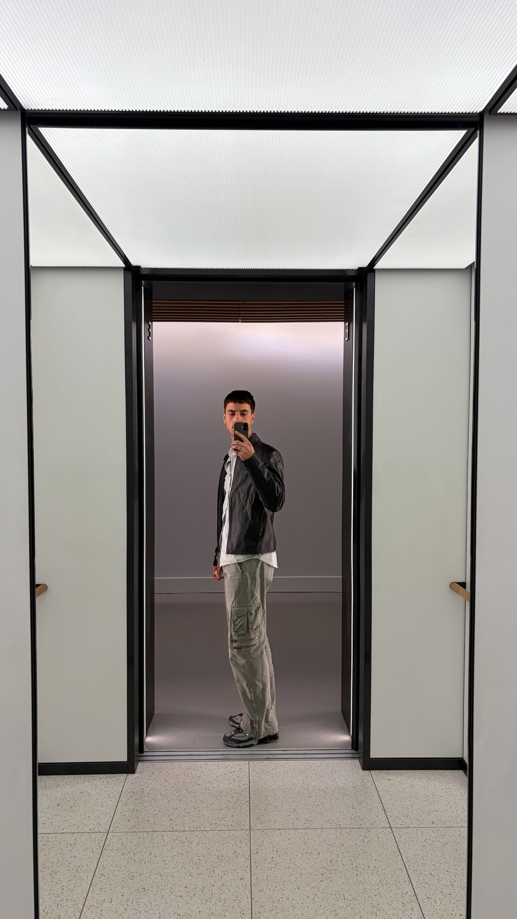 A man takes a selfie in an elevator, wearing a black leather jacket over a white t-shirt and light-colored pants.