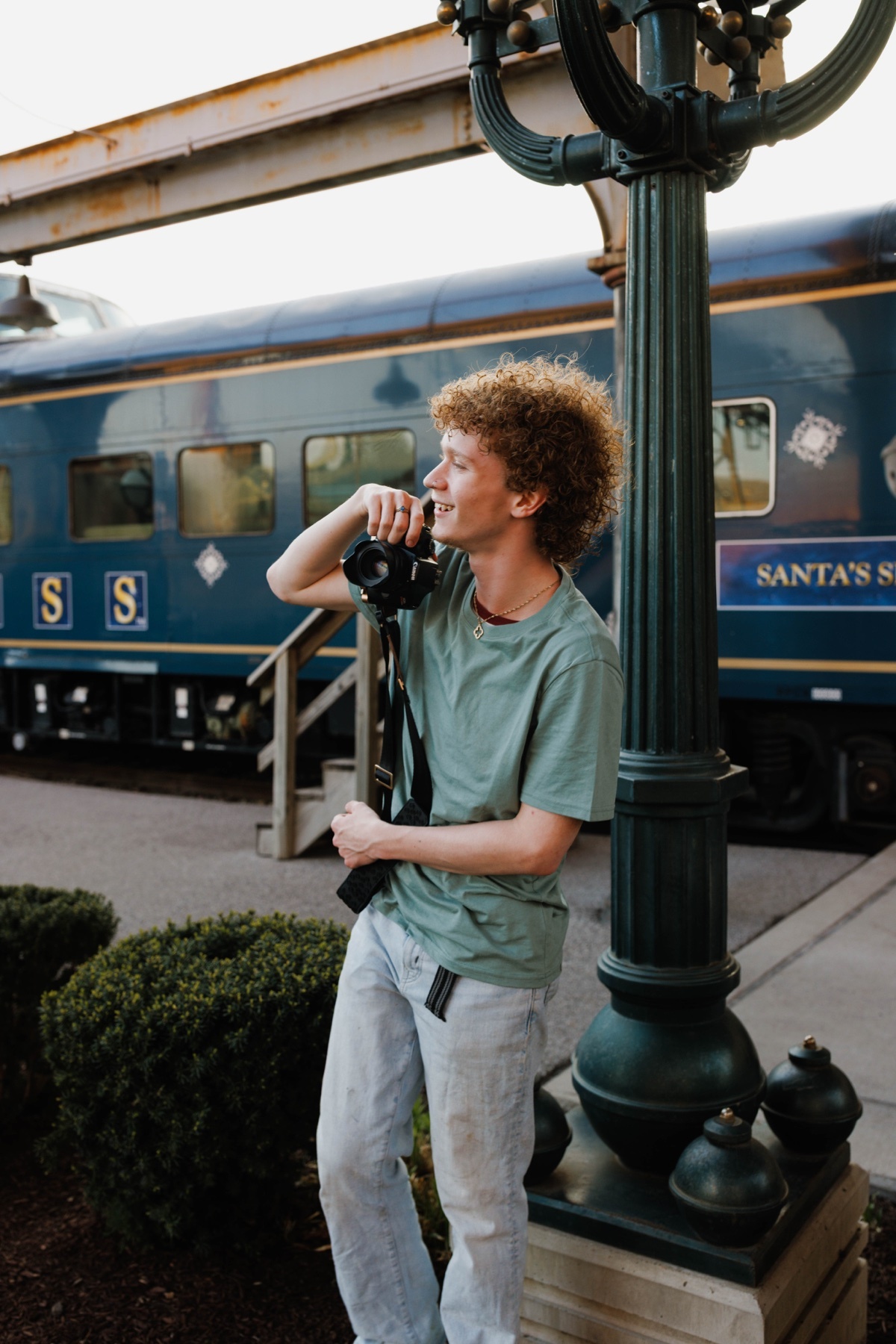 A young man with curly hair holds a camera and poses next to a vintage train. He is wearing a light green t-shirt and light-colored pants.