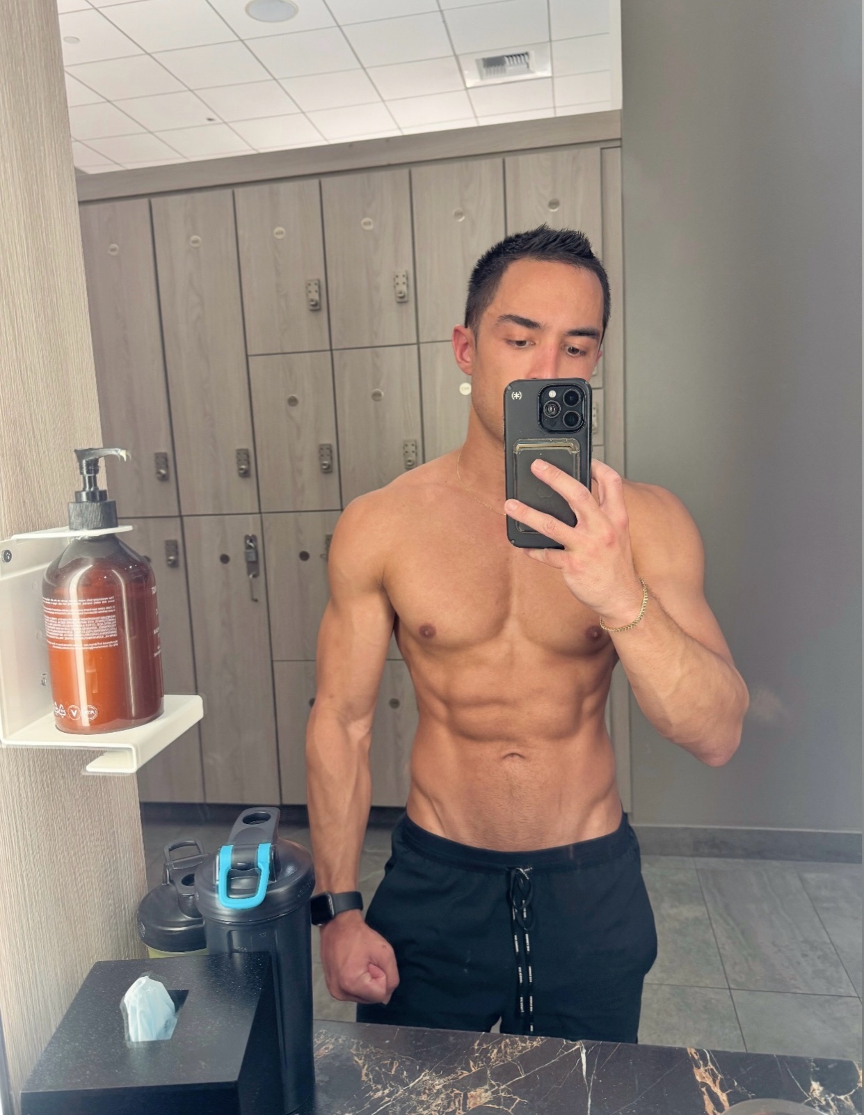 A man takes a mirror selfie in a gym locker room, showing off his muscular physique and abs. He is wearing black shorts and is shirtless.