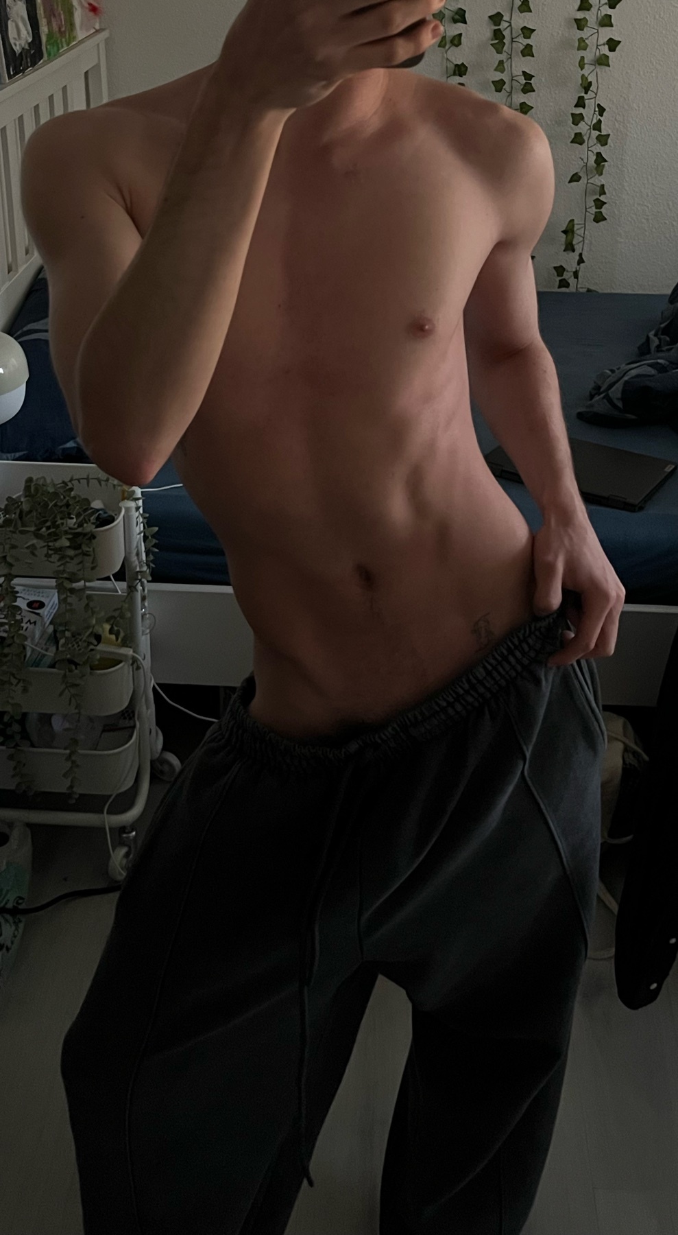 A shirtless man in sweatpants poses in a bedroom, showing off his abs and athletic physique.