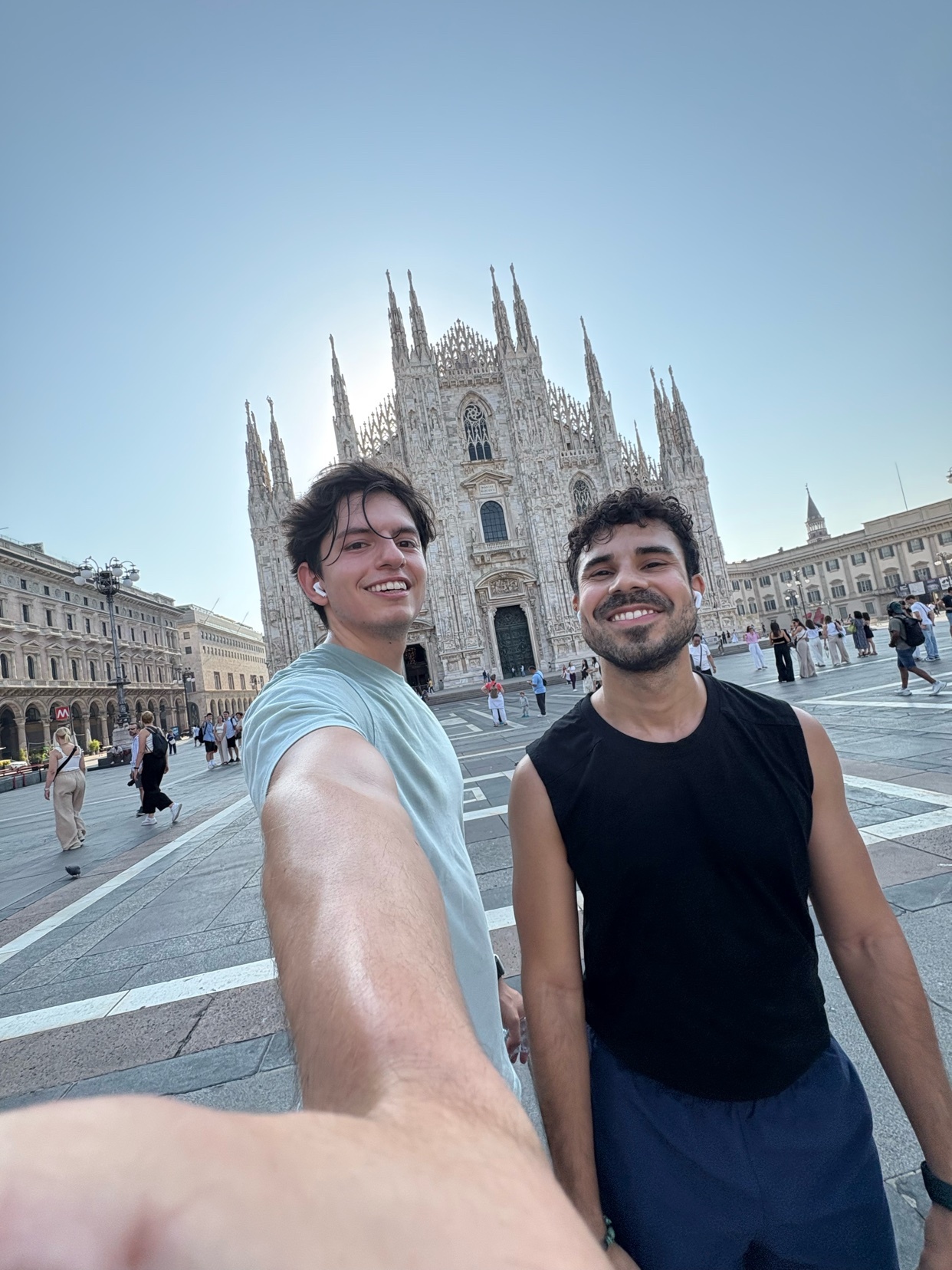Two men smile for a selfie in front of the Duomo di Milano. The iconic cathedral stands tall in the background on a sunny day.