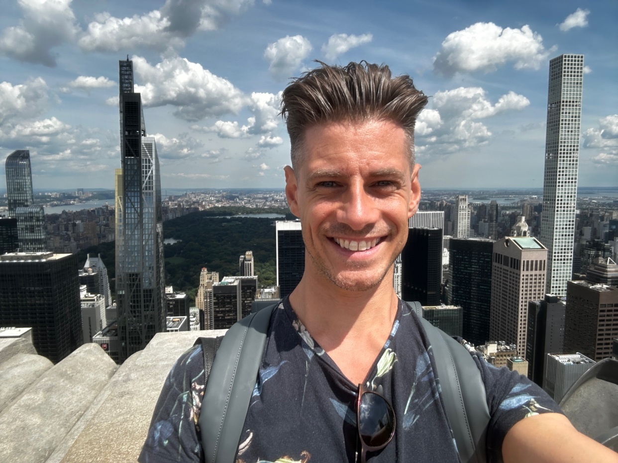 A man takes a selfie with a stunning cityscape in the background. The photo captures a vibrant day with a clear sky and modern buildings.