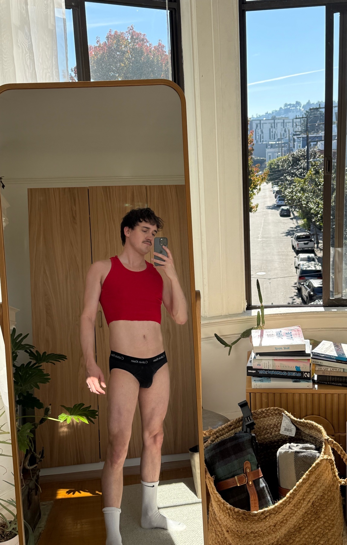A man poses in front of a mirror, wearing a red crop top and underwear. The photo is taken in a well-lit room with a view of a street outside the window.