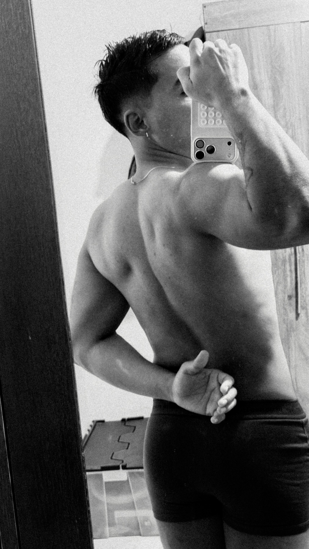 A man takes a mirror selfie, showcasing his physique. The black and white image highlights his muscular build and shirtless torso.
