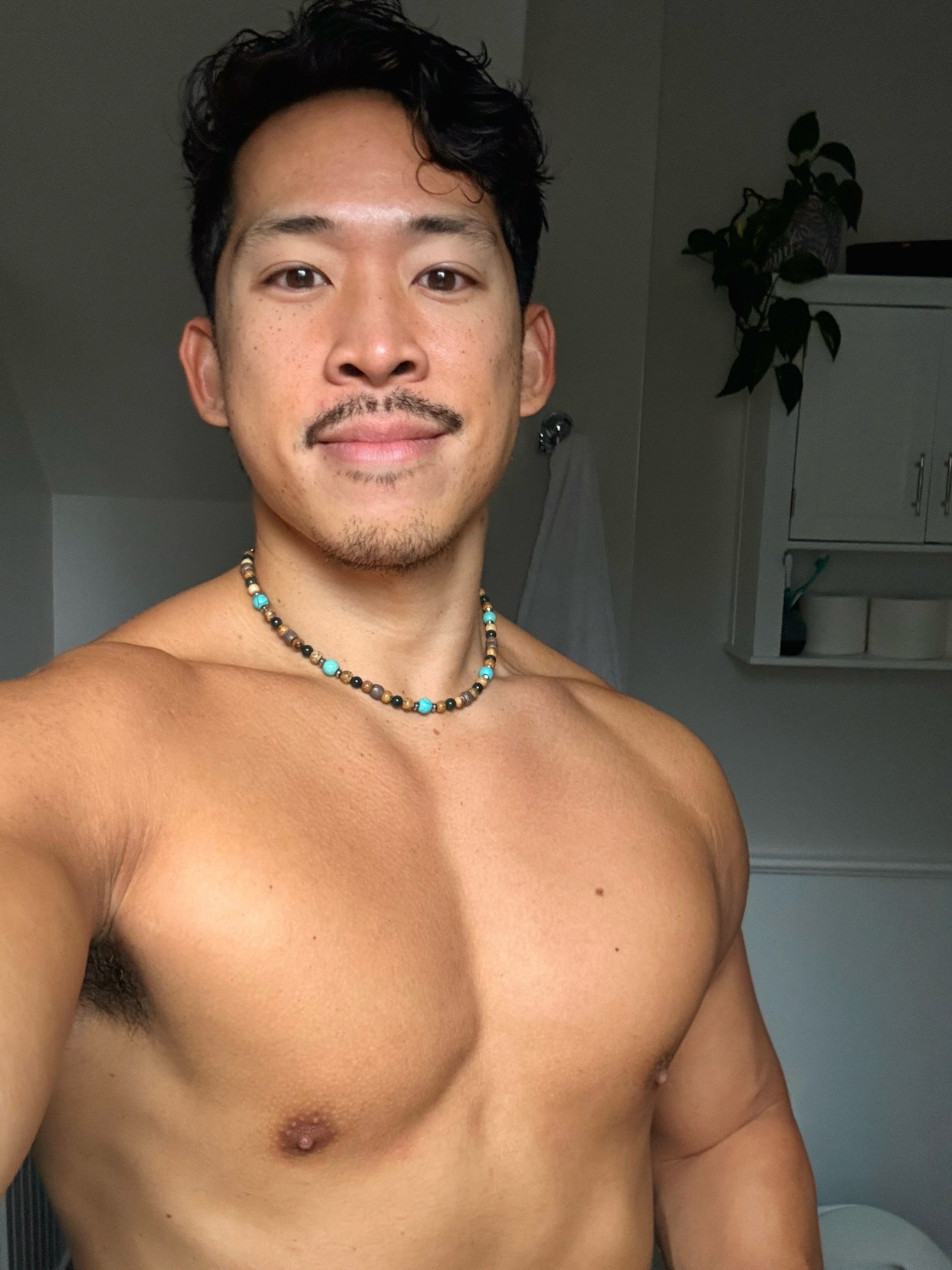 A shirtless man with dark hair and a mustache poses for a selfie. He is wearing a necklace and has a muscular physique.