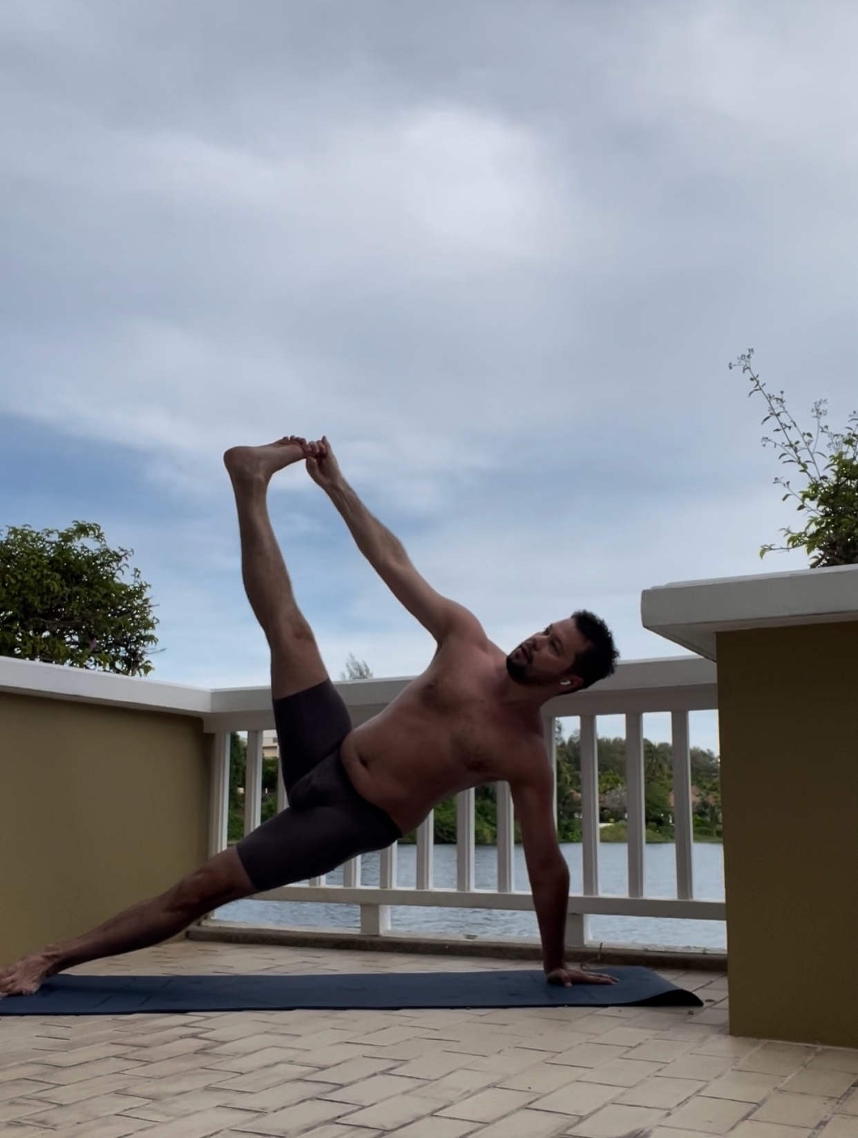 A man is doing yoga on a mat outdoors. He is shirtless and has a muscular physique.