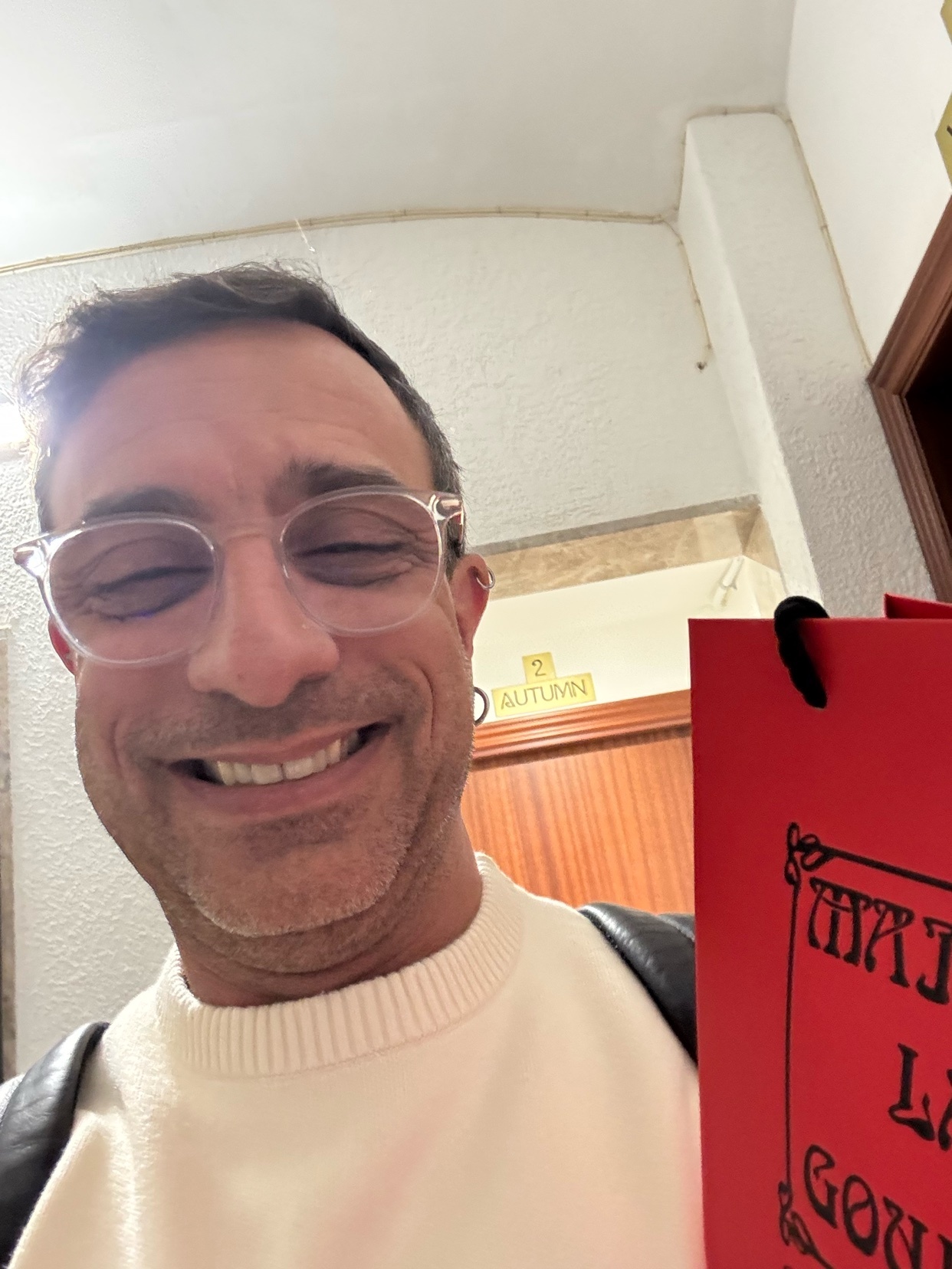 A man smiles at the camera while holding a shopping bag. He is wearing glasses and a white sweater.