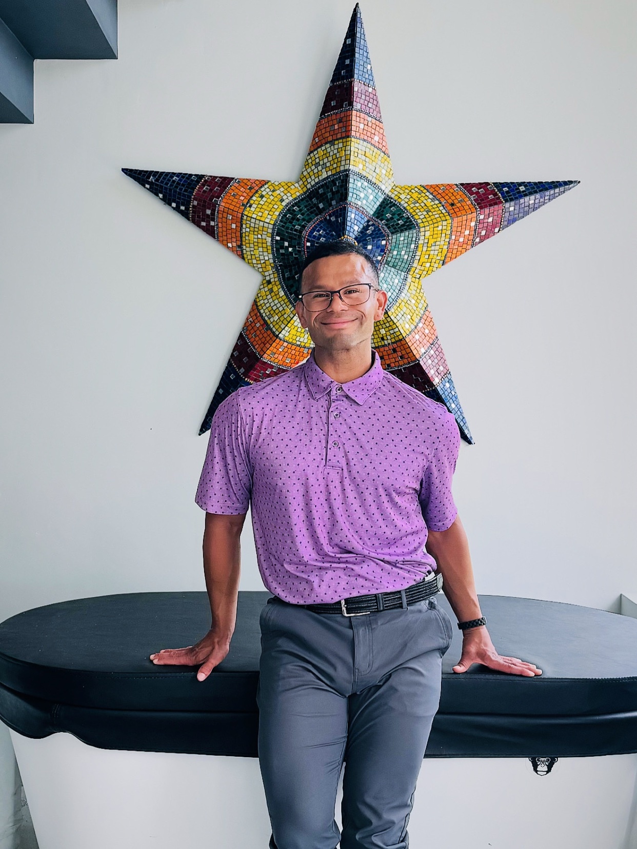 A man poses in front of a colorful star-shaped artwork. He is wearing a purple shirt and glasses.