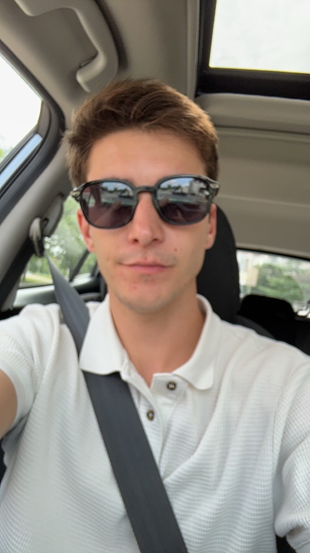 A young man wearing sunglasses and a white polo shirt takes a selfie while driving in a car.