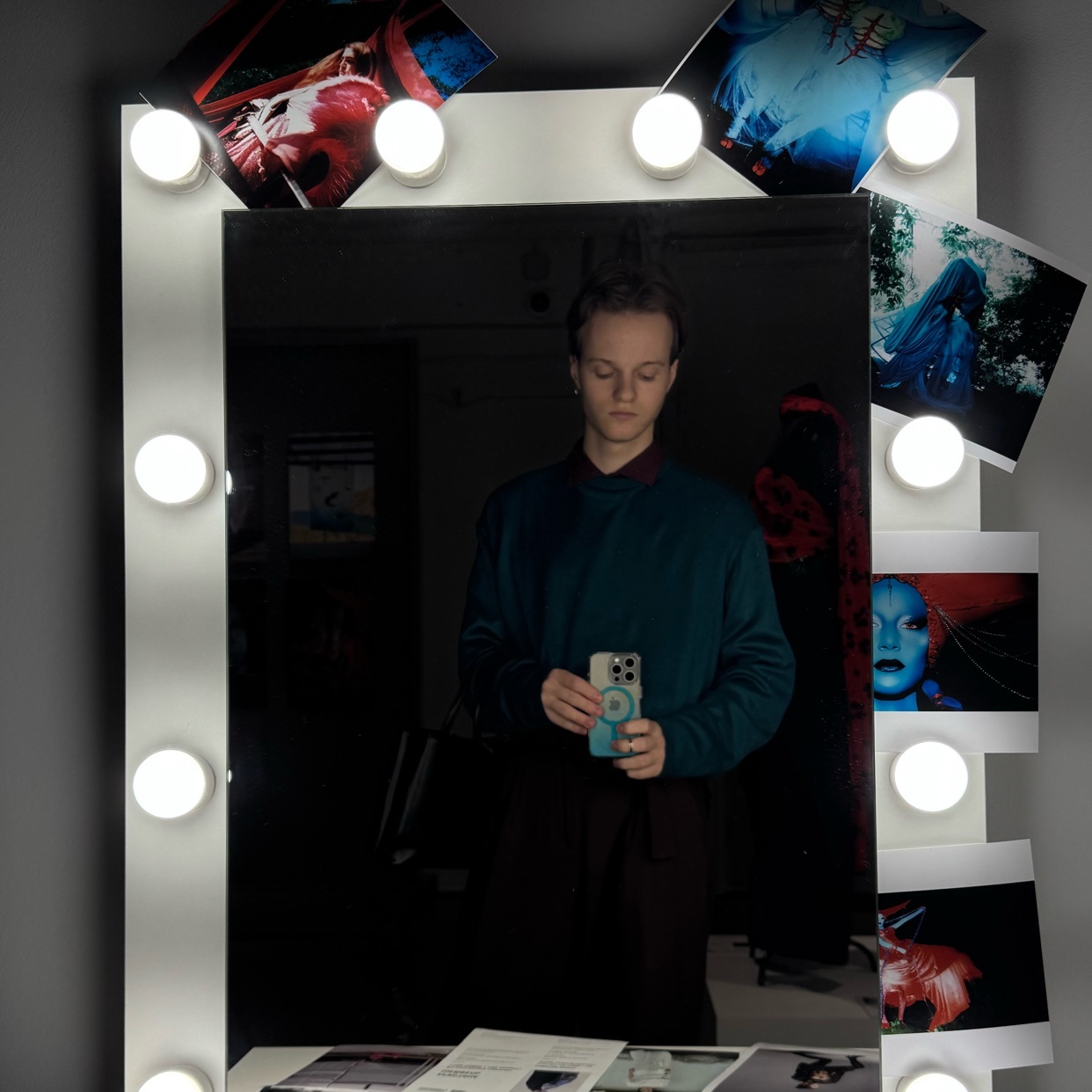 A man takes a mirror selfie in a stylish outfit. The mirror is surrounded by lights and photos.